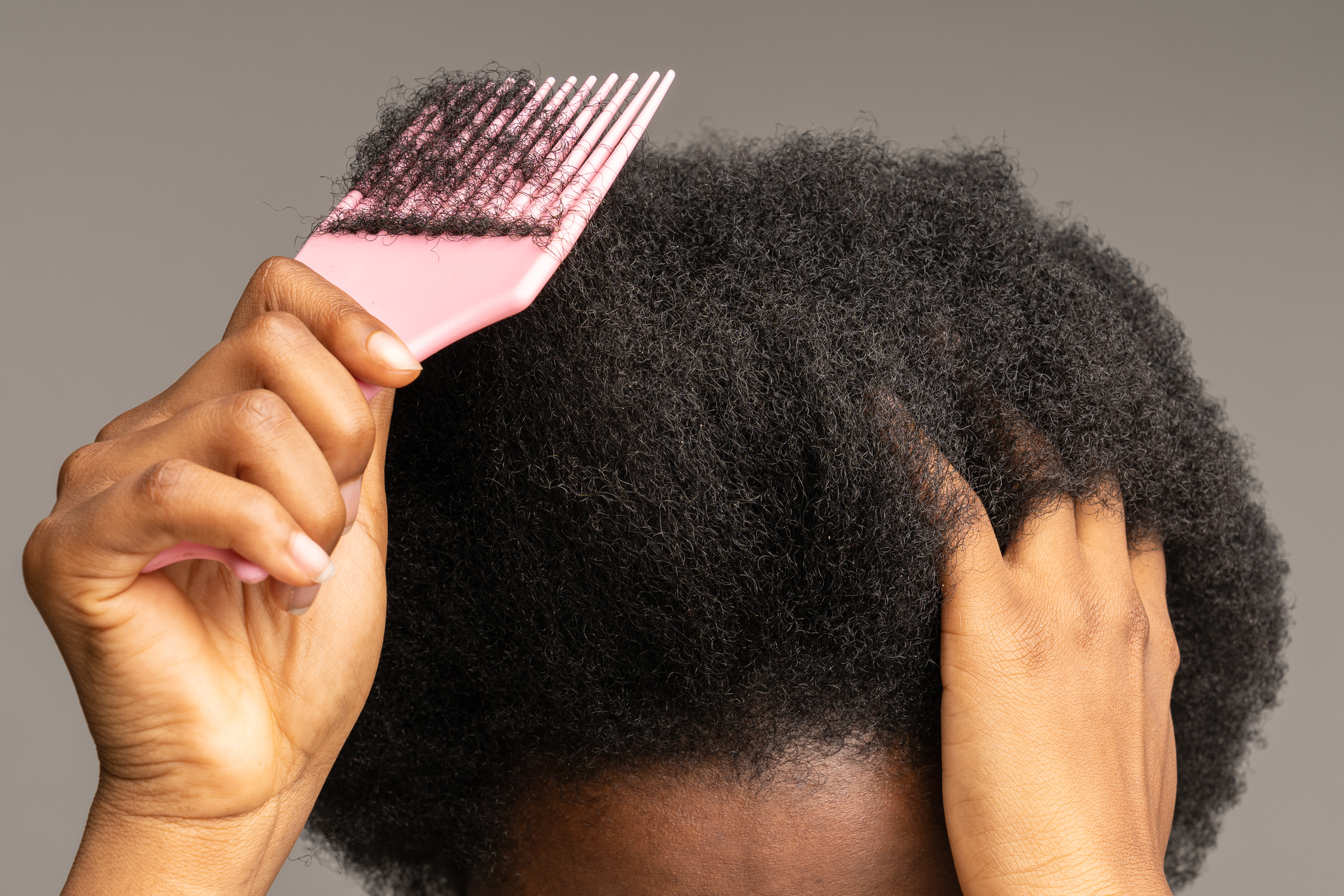 Close up of African female using comb, brushing curly trick hair isolated on studio gray wall.